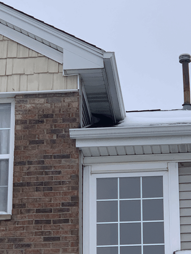 A dark gap beneath the roof soffit and return, positioned directly above a snow-covered gutter intersection on a residential home.
